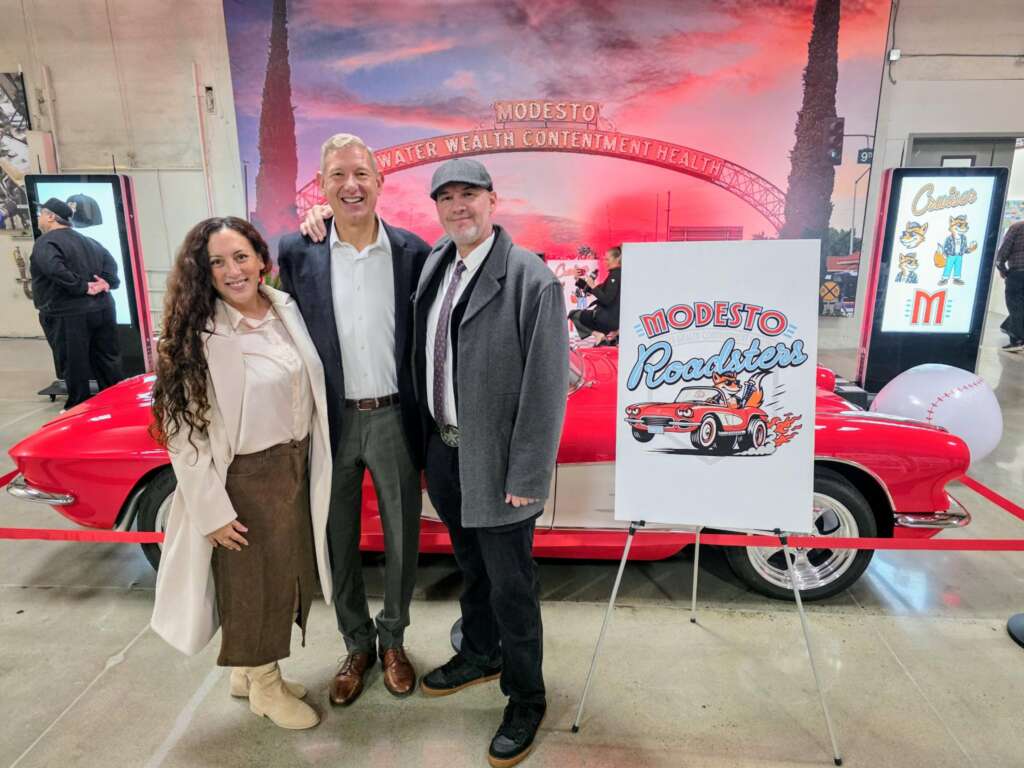 Three guests beside the Modesto Roadsters logo board and the red Corvette, under the Modesto arch mural at the reveal