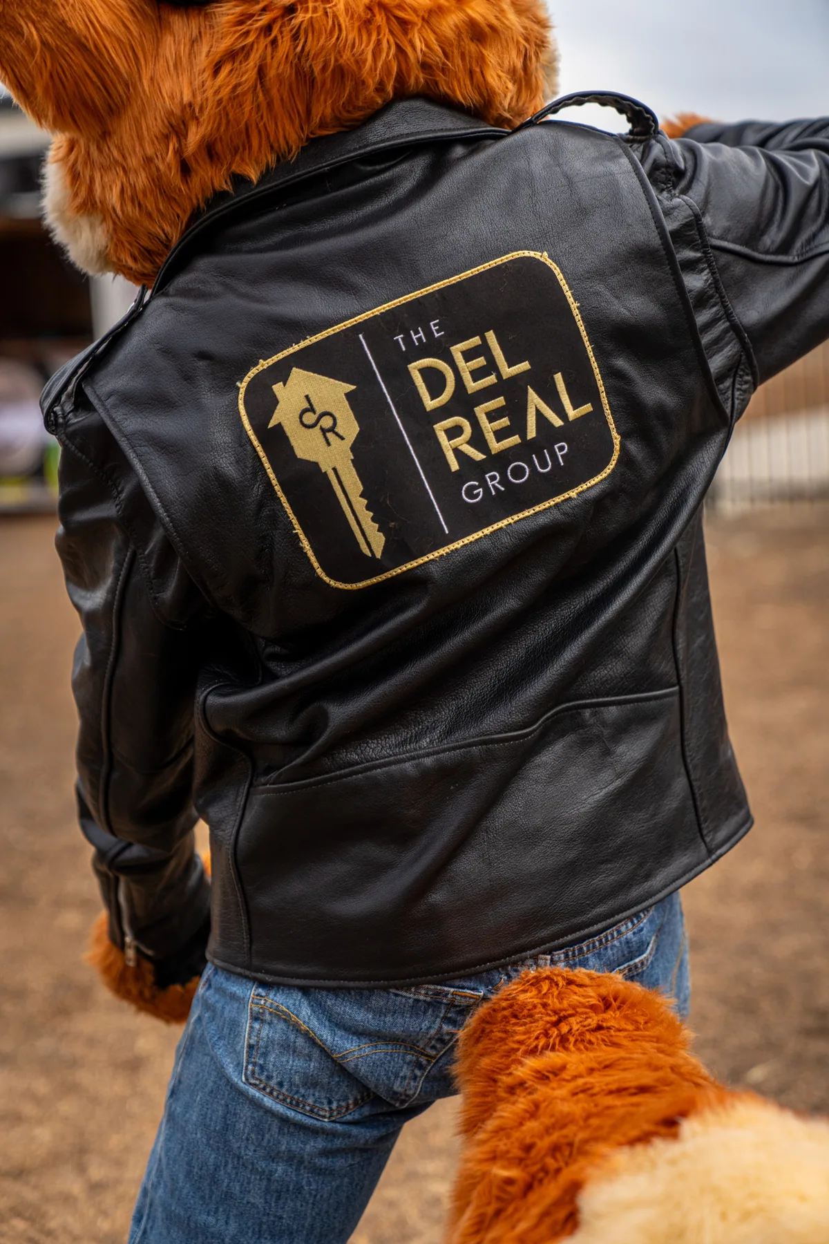 The back of Cruiser's black leather jacket, stitched with THE DEL REAL GROUP and the company's gold house-and-key mark