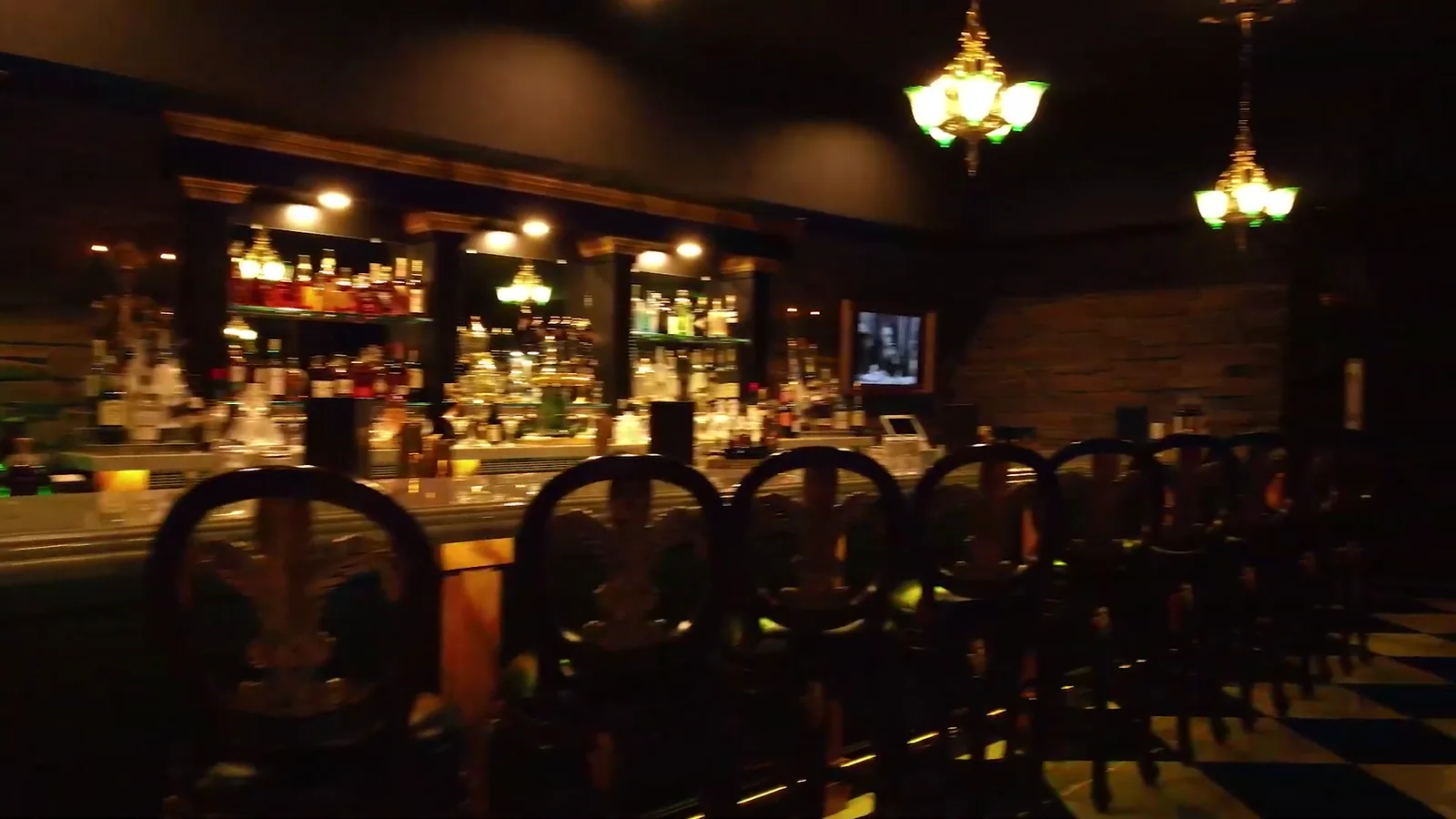 The Falcon's Lair back bar at night: bottles lit along a mirrored wall, two chandeliers above, arched chair backs in the foreground