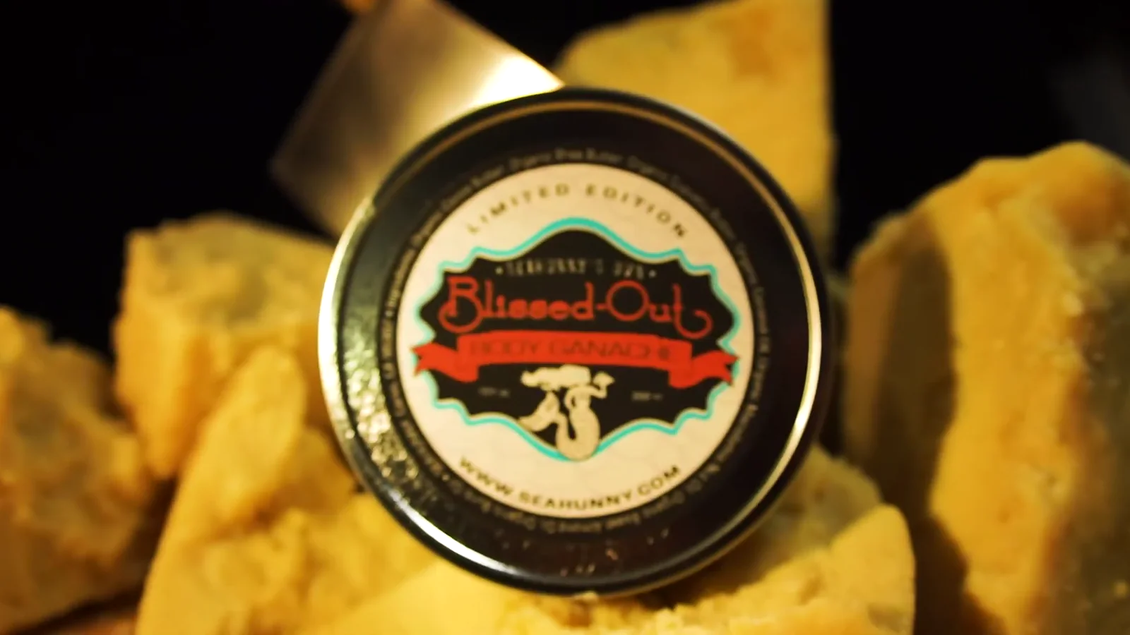 A finished Seahunny Blissed-Out Body Ganache tin, gold lid facing the camera, surrounded by broken chunks of raw shea butter