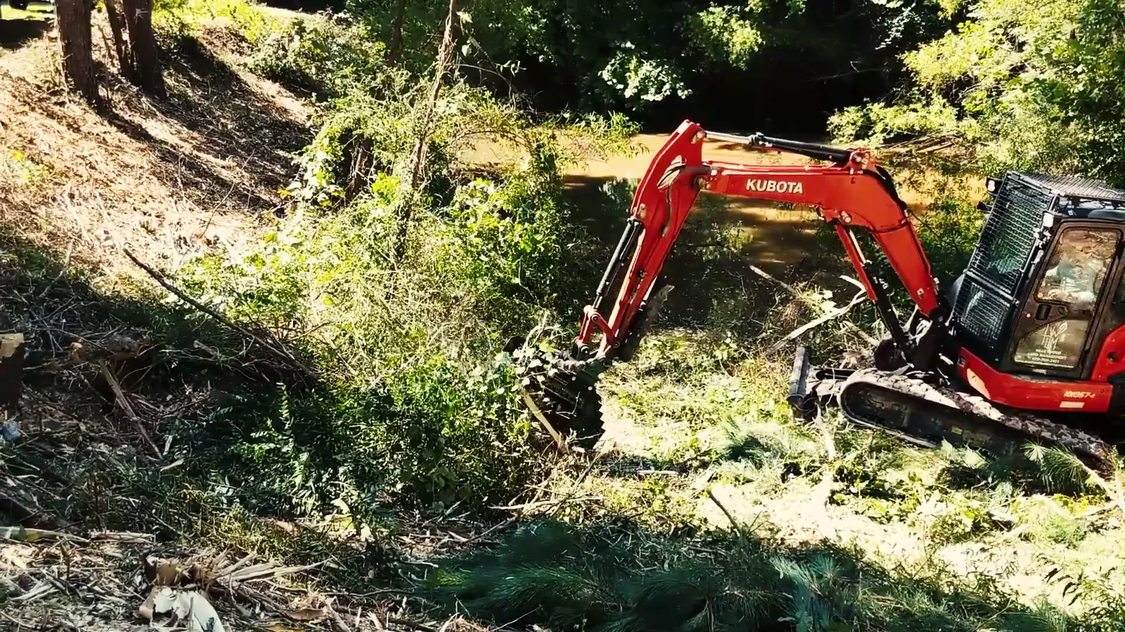 A red Kubota excavator with a BrushHound head clearing brush from a steep pond bank, cut material spread across the slope