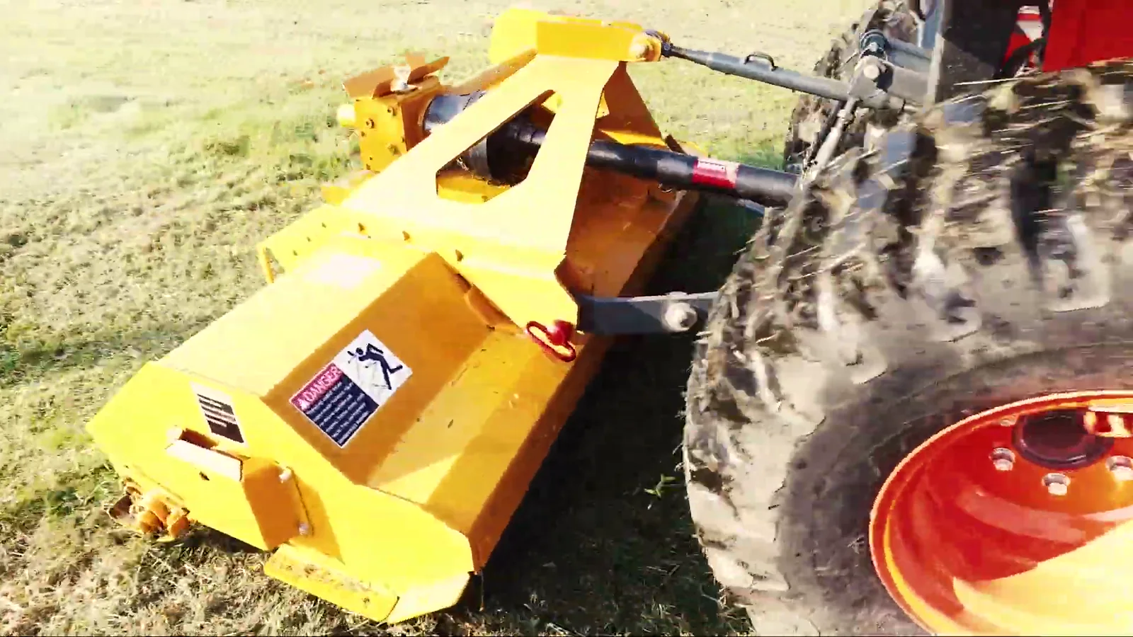 Close view of the yellow Vrisimo MiniMax flail mower hitched behind the rear wheel of a red compact tractor on fallen autumn leaves