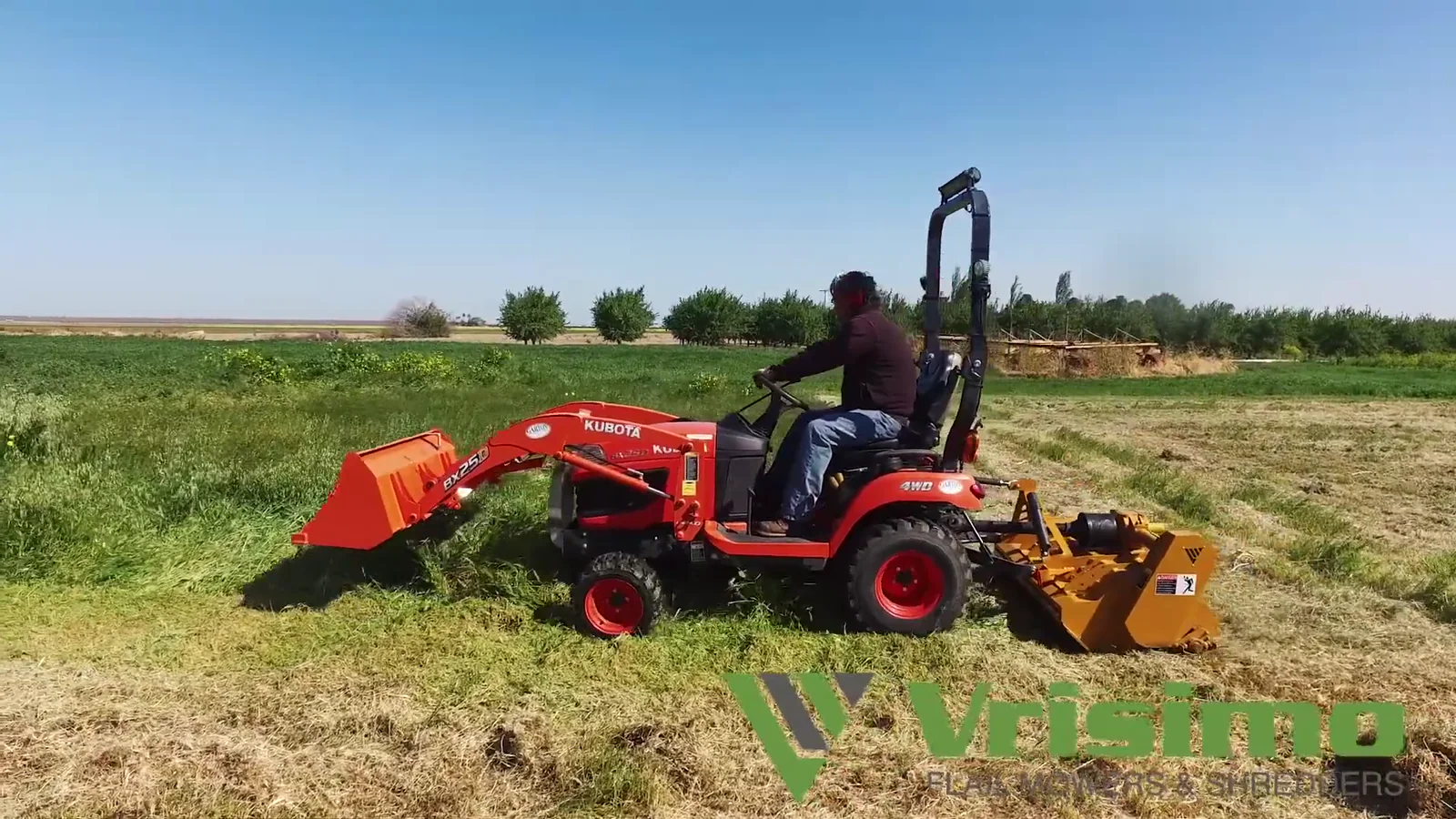A compact orange tractor towing a yellow Vrisimo MiniMax flail mower through open green field under a clear sky, a cut swath behind it