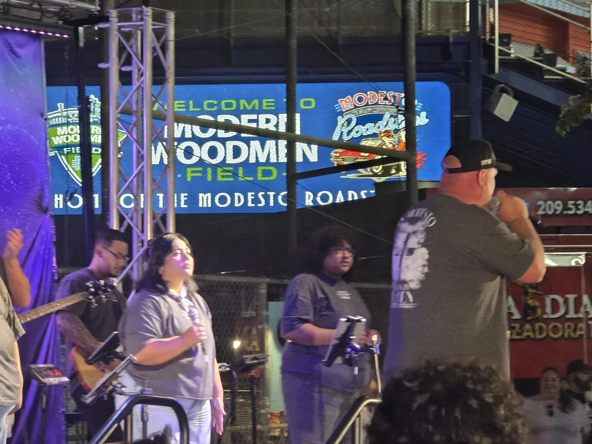 Modesto Downtown Worship performing the postgame Faith Fest set at Modern Woodmen Field: singers, a bass player and a speaker on stage beneath the Welcome to Modern Woodmen Field sign