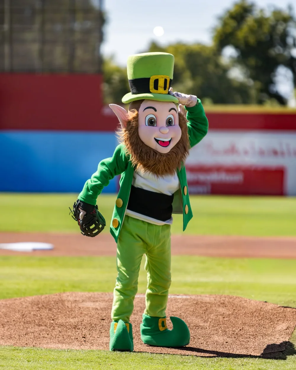 Rally O'Malley on the pitching mound in a green coat, waistcoat and buckled top hat, tipping the hat with a fielding glove on the other hand