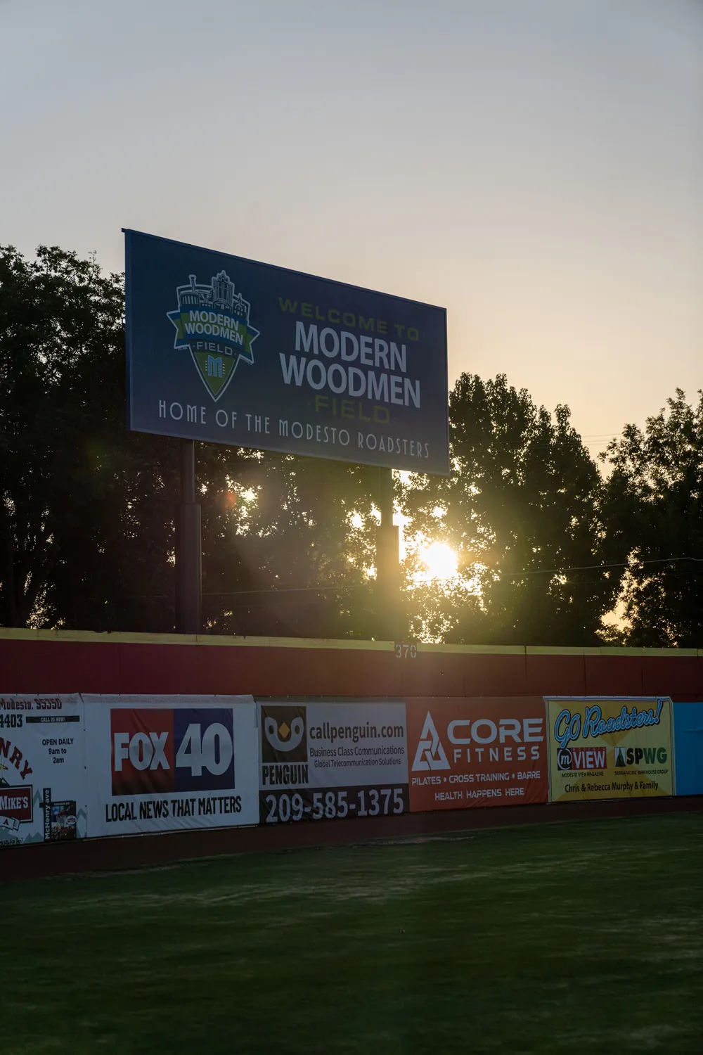 Late afternoon sun coming through the trees behind the outfield sign.